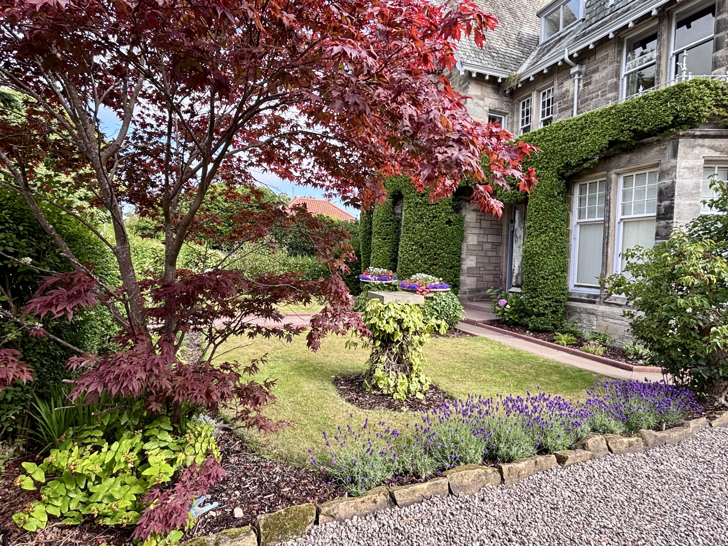 Flower beds and a red-leaved tree beside the stone facade